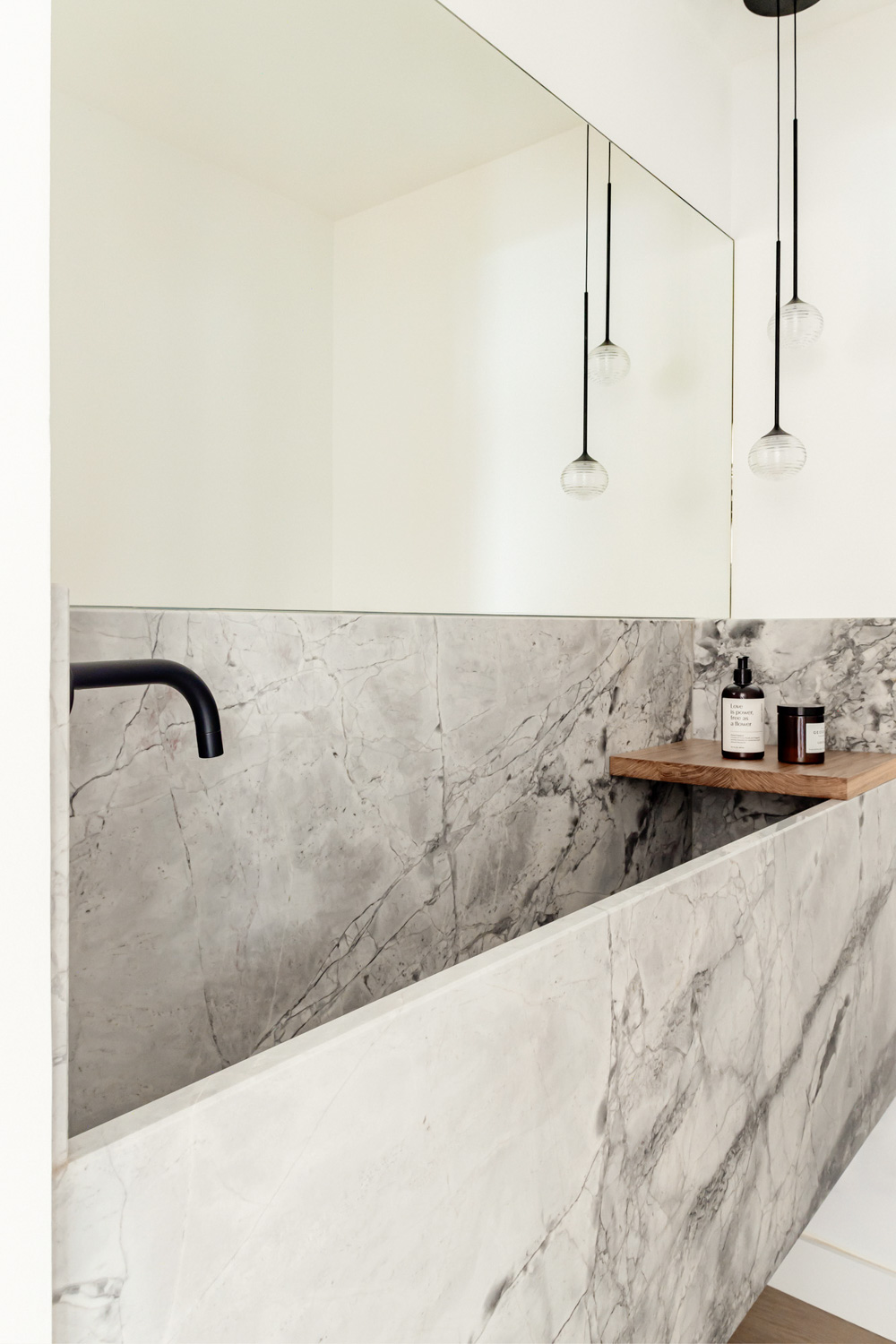 ABATON-reforma-en-el-escorial-1 Bañera moderna de mármol gris con grifería negra mate y estantería de madera, iluminación colgante minimalista y espejo de gran formato