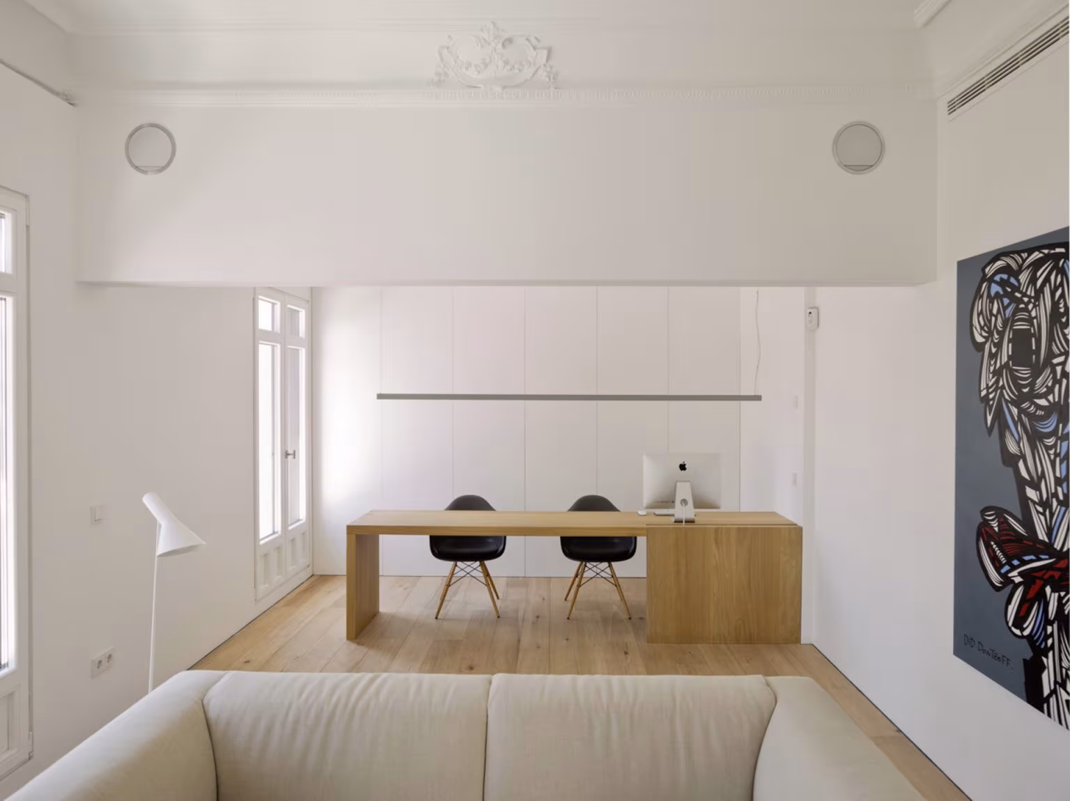 Sala con mesa de trabajo de roble y sillas negras entre muros blancos y un sofá en primer plano, en Alcalá
