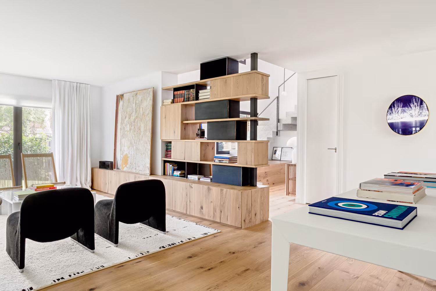 Salón con estantería de roble y metal negro como separador, butacas negras y suelo de madera en Paseo de Alcobendas