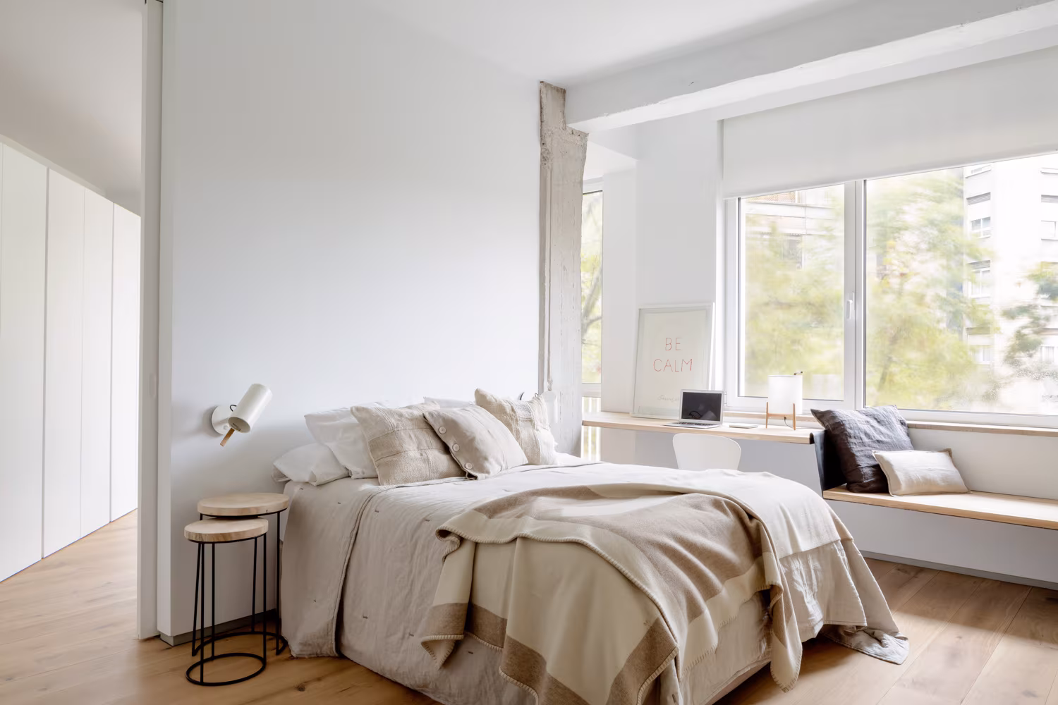 Dormitorio con ropa de cama clara, banco bajo la ventana y pilar de hormigón visto, en Santiago Bernabéu