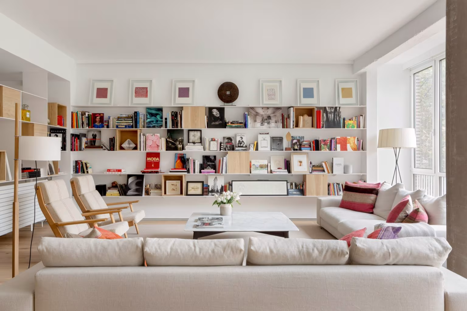 Living room with a continuous white bookcase, pale sofa and wooden armchairs in Ábaton's Santiago Bernabéu refurbishment