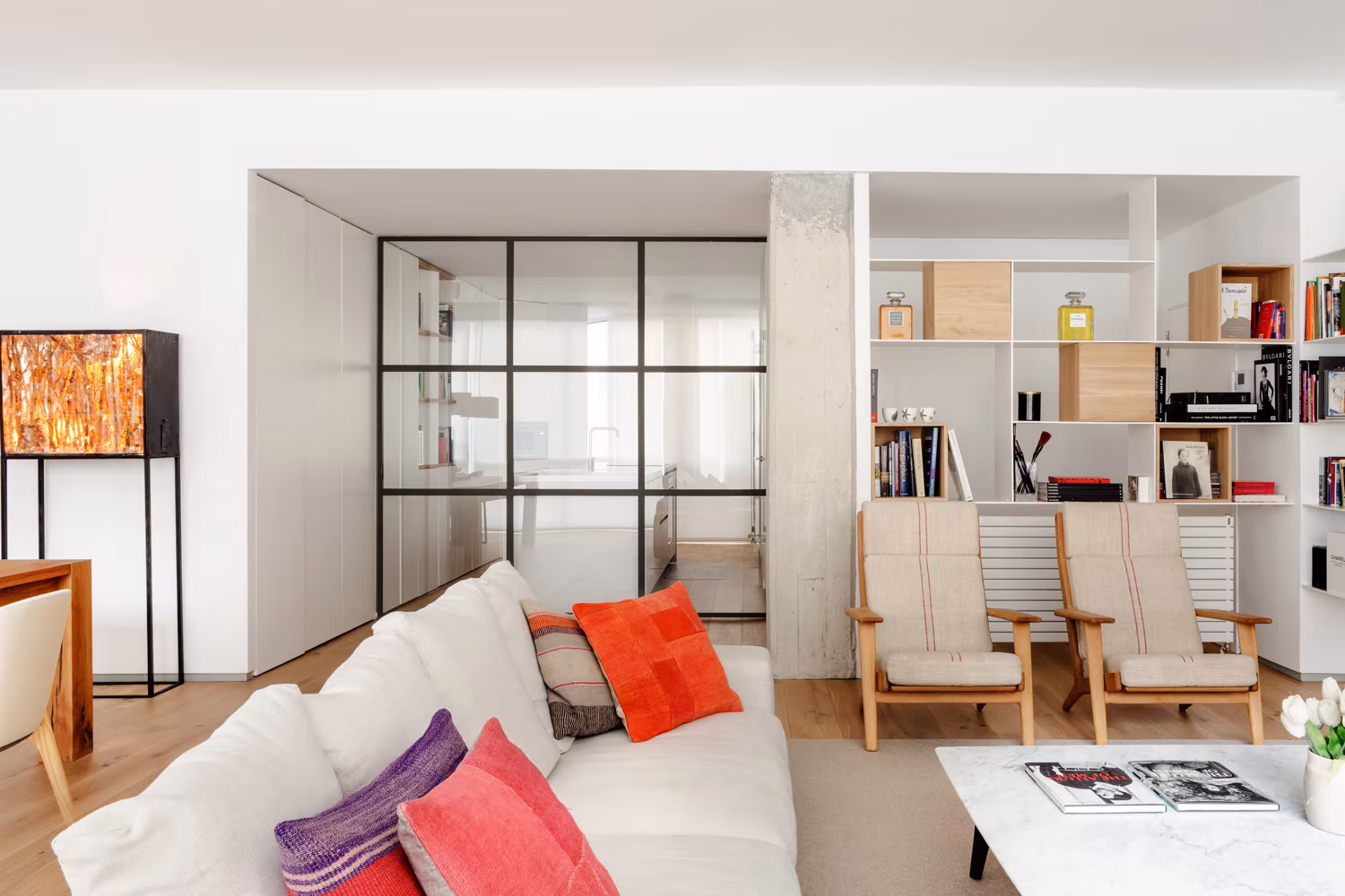 Sala con cerramiento de vidrio y acero, sofá blanco con cojines de colores y librería, en la reforma de Santiago Bernabéu