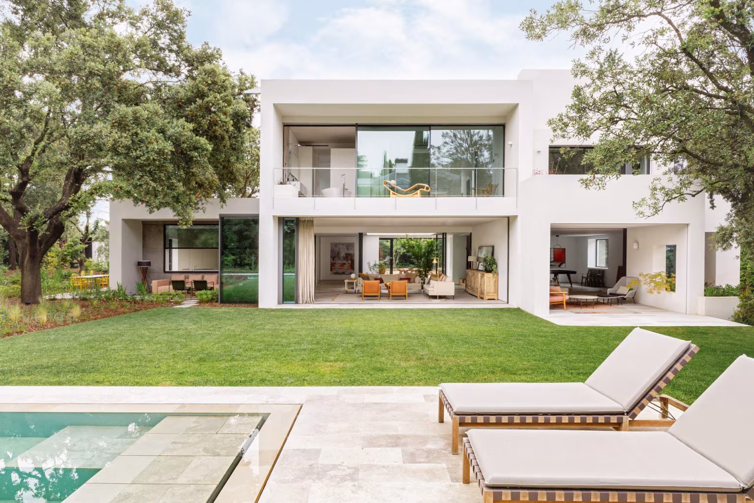 Casa L de Ábaton con dos plantas blancas, césped, tumbonas y piscina entre encinas