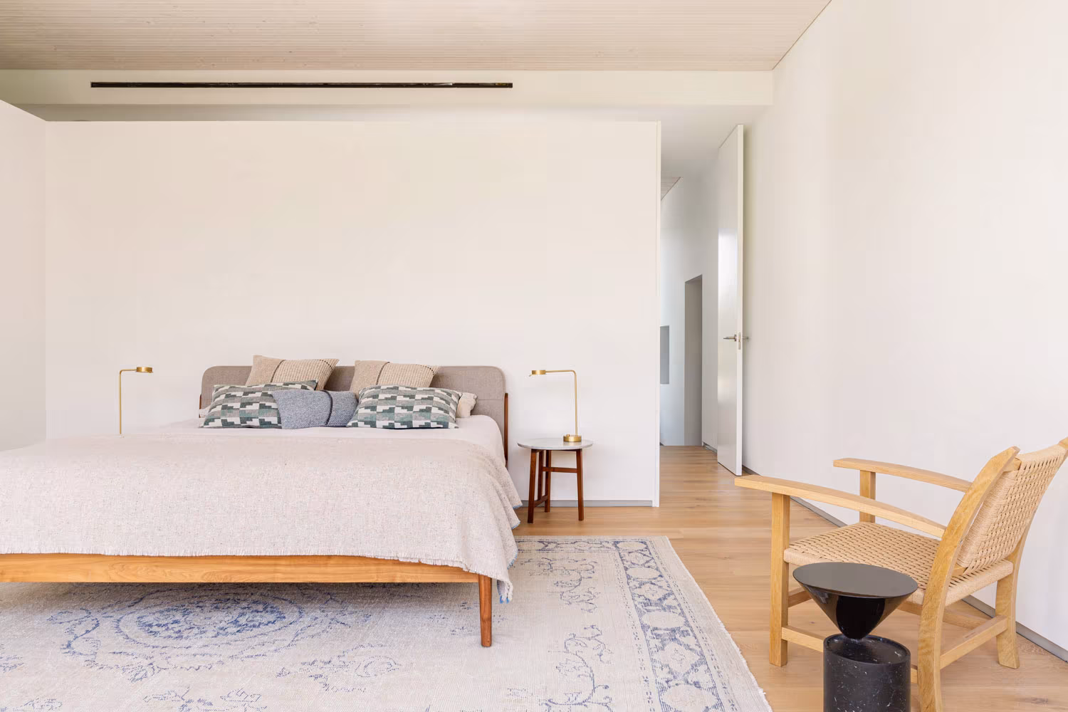 Bedroom with timber bed, pale rug and cane armchair before a white volume, LVL refurbishment