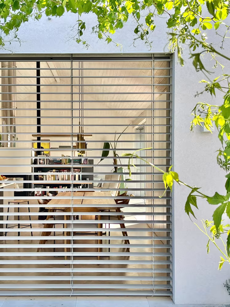 Window with horizontal slats among green leaves, a work table visible behind