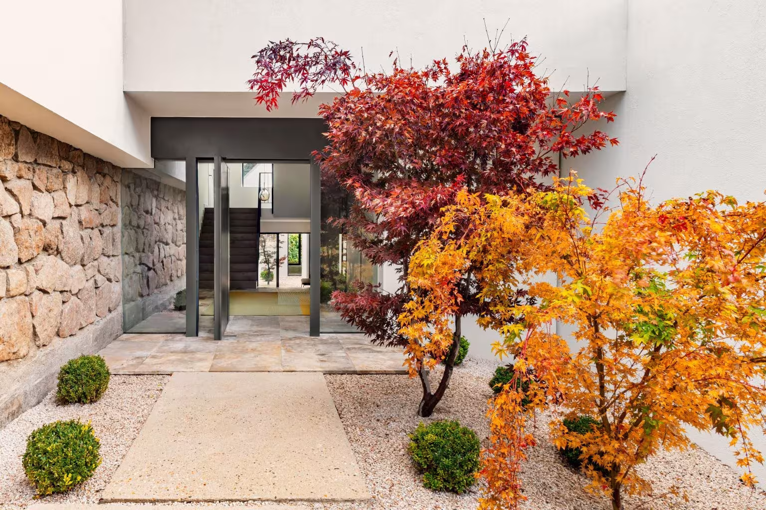 Entrada de casa moderna con jardín de grava, árboles japoneses en otoño y fachada de piedra natural.