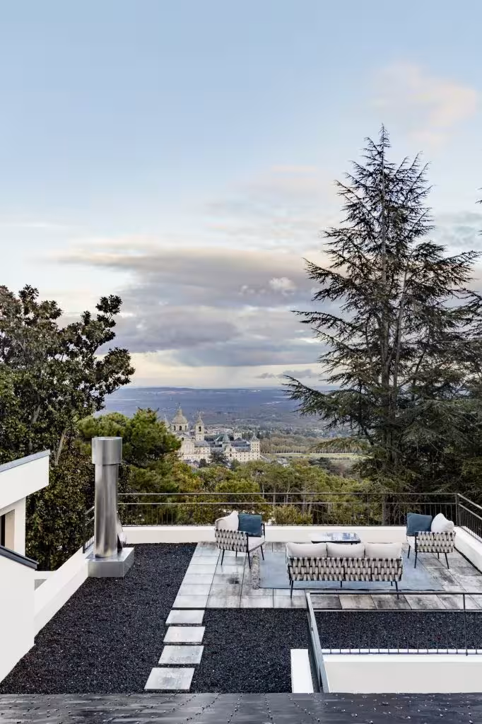 Terraza con zona de estar moderna, pavimento de piedra y vistas espectaculares al Monasterio de El Escorial entre pinos.