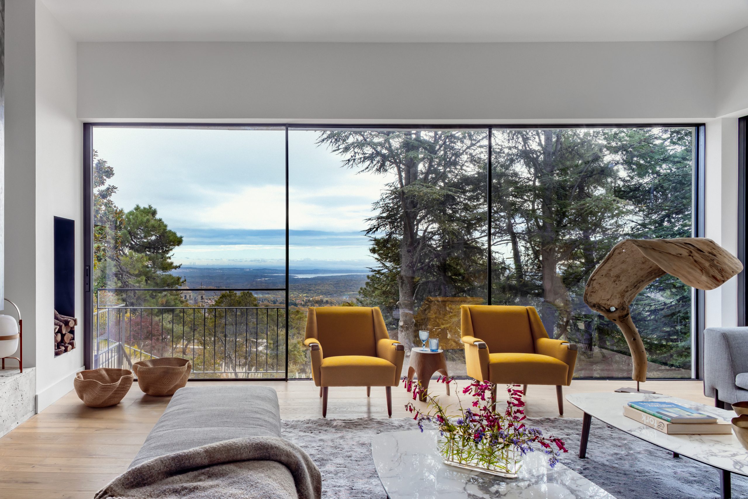 EL ESCORIAL- 3 Salón moderno con grandes ventanales, dos sillones mostaza, vistas panorámicas al bosque y mesa de centro con flores