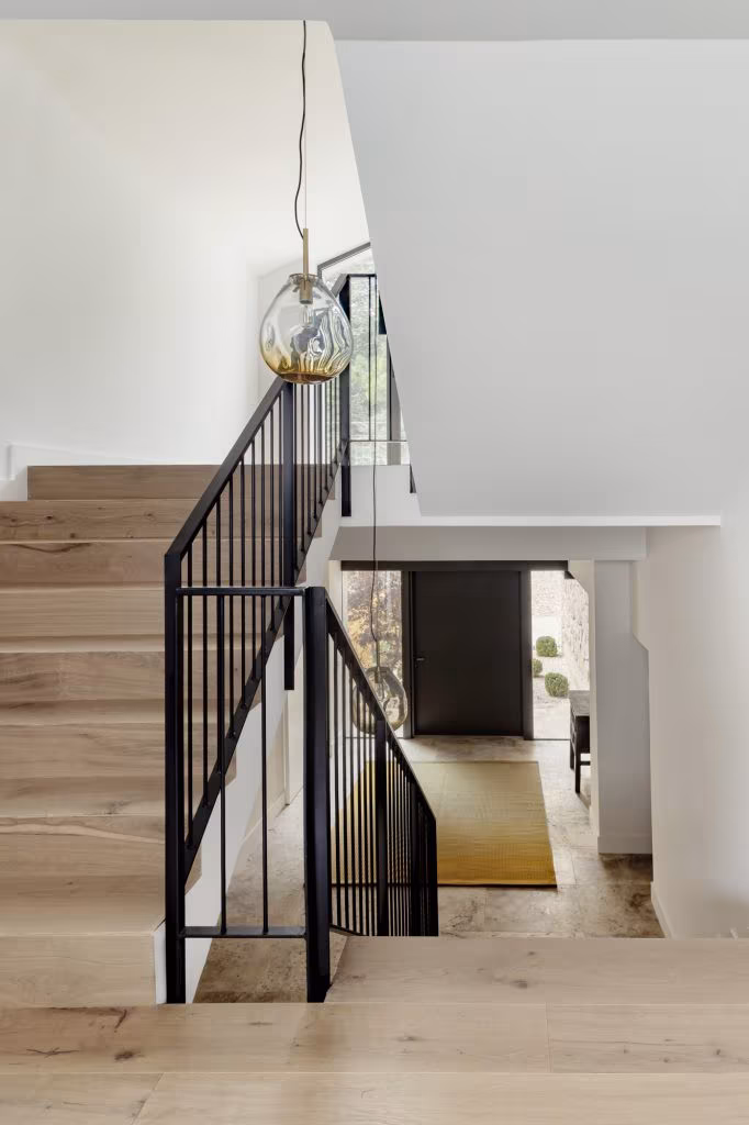 Escalera interior de madera con barandilla metálica negra, lámpara colgante de diseño y recibidor con alfombra amarilla y gran puerta negra