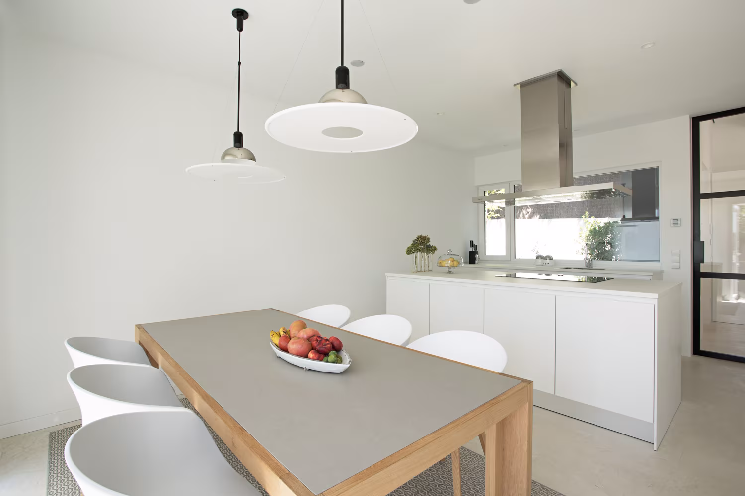Cocina blanca con mesa de madera y encimera gris, lámparas colgantes y campana de acero en Casa JP de Ábaton