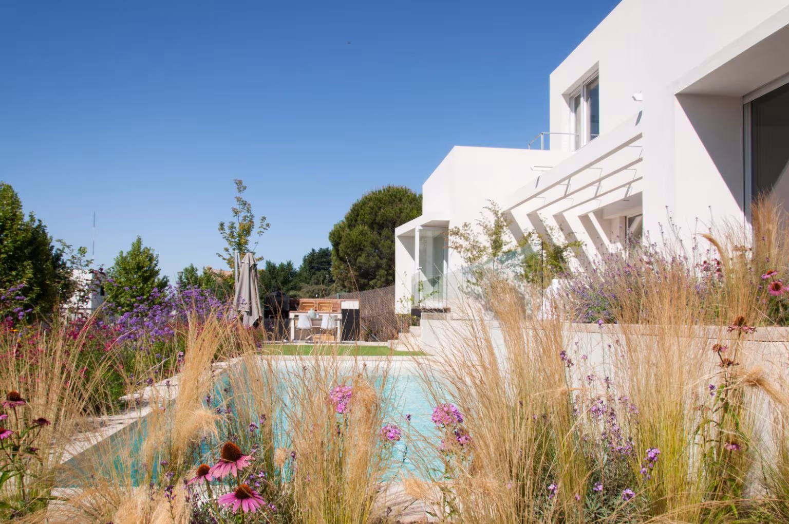Casa JP de Ábaton con piscina rodeada de gramíneas y flores bajo una fachada blanca escalonada, Madrid