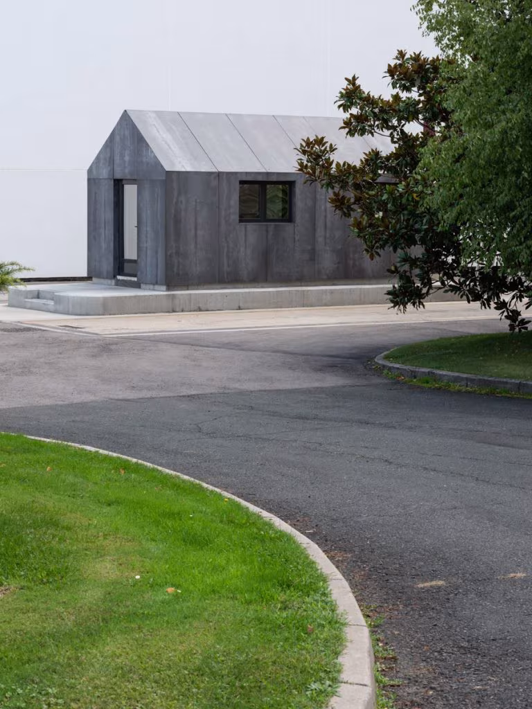 Small dark concrete pavilion with a metal gabled roof beside a path and a magnolia at CVNE