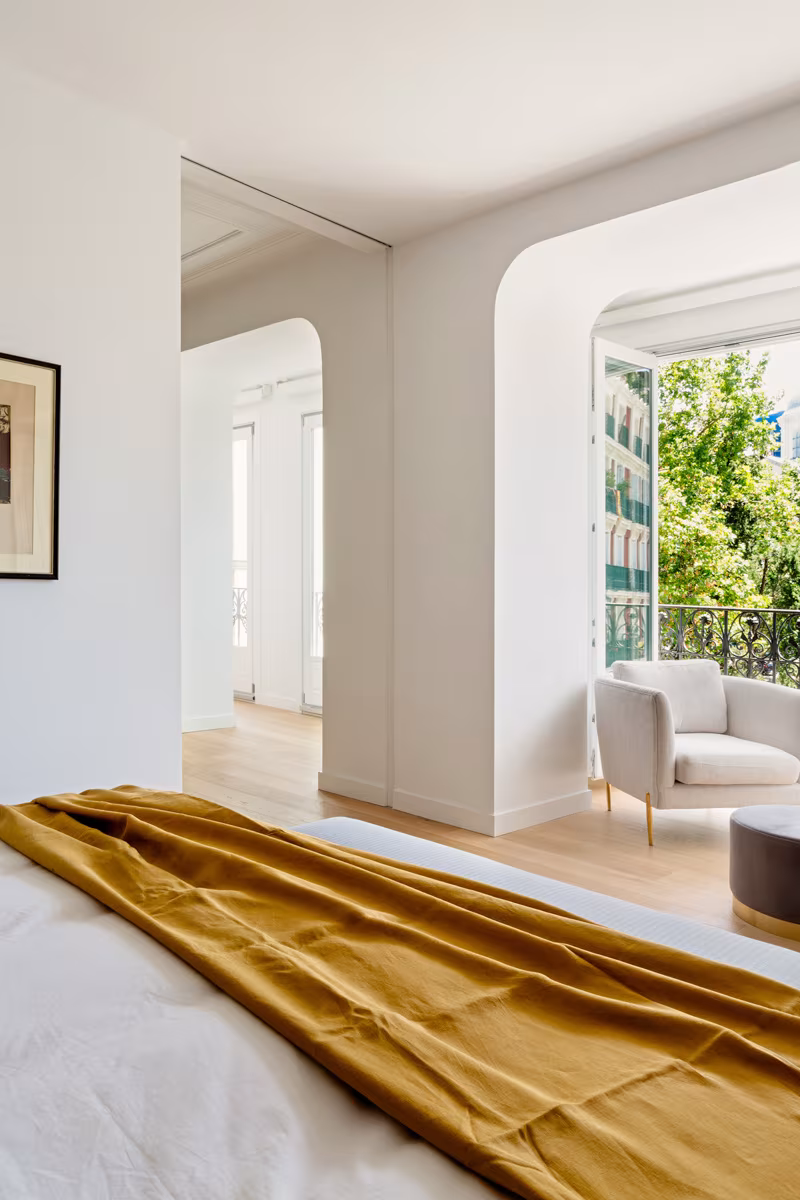 Cama con manta mostaza ante un mirador arqueado con vistas a los árboles en la reforma de Salesas