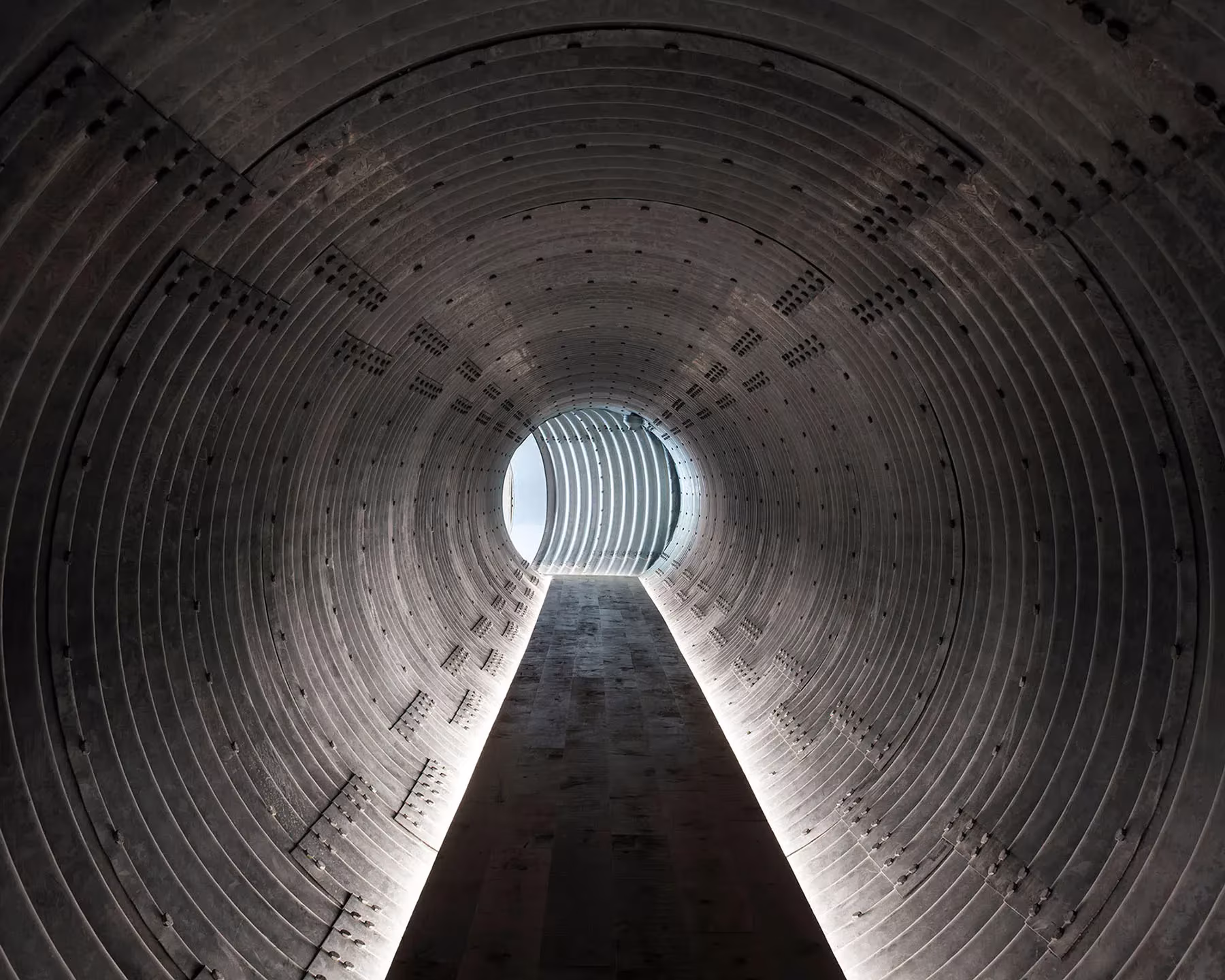 Túnel circular de chapa metálica con una franja de luz en el suelo y una apertura al fondo, en el pabellón enterrado