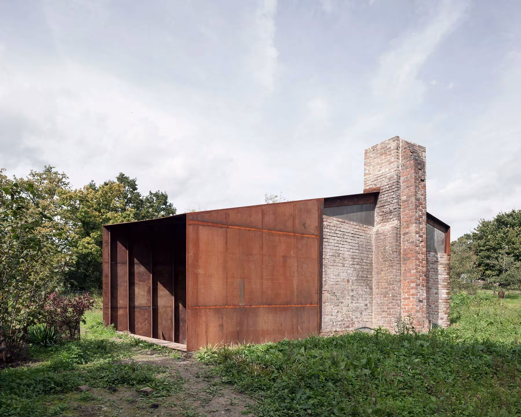 Esquina de acero corten del pabellón de Carmody Groarke junto a la chimenea de ladrillo rehabilitada