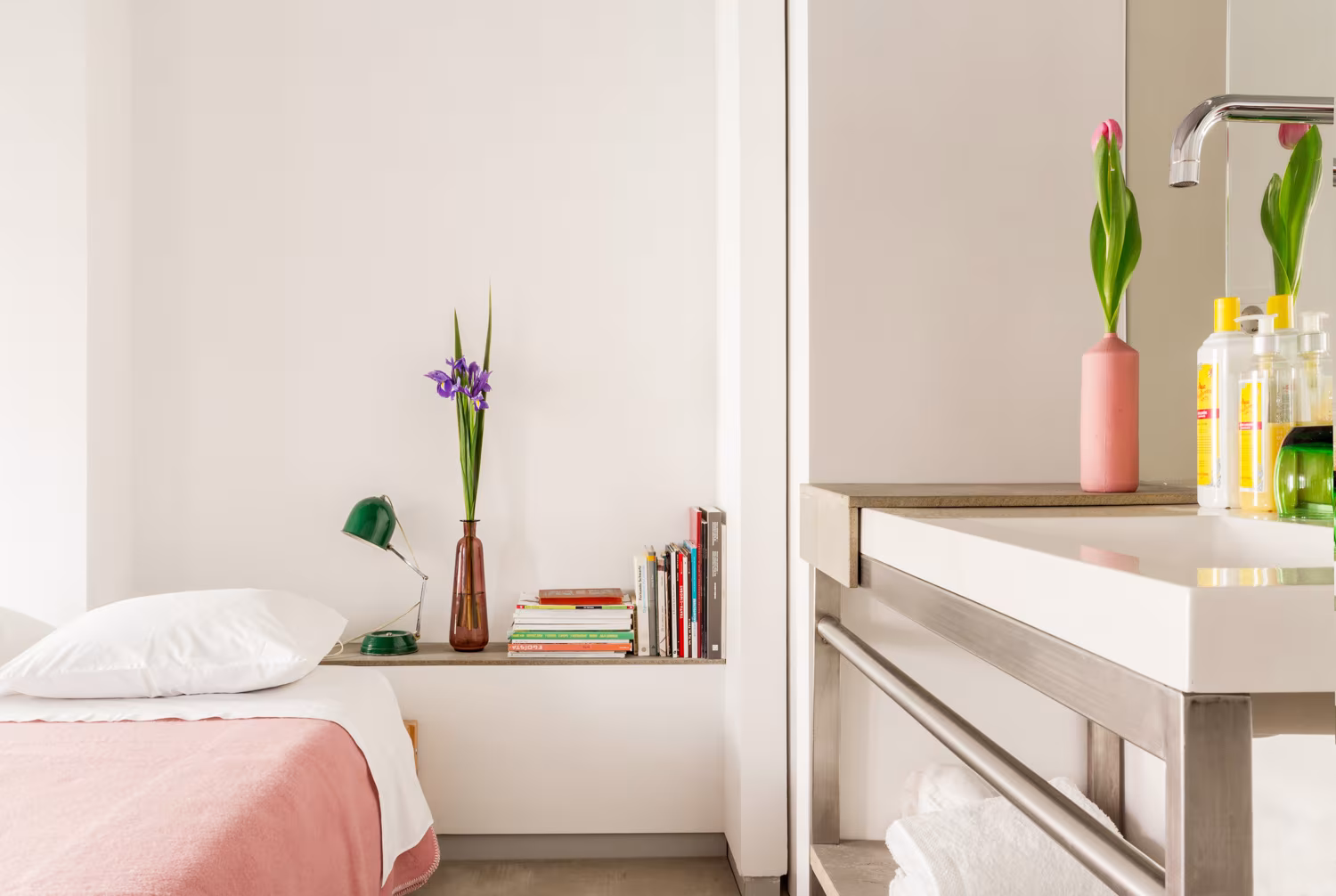 Cama con manta rosa, repisa con libros y flores y lavabo de acero junto a la cabecera en el alojamiento de Lisboa
