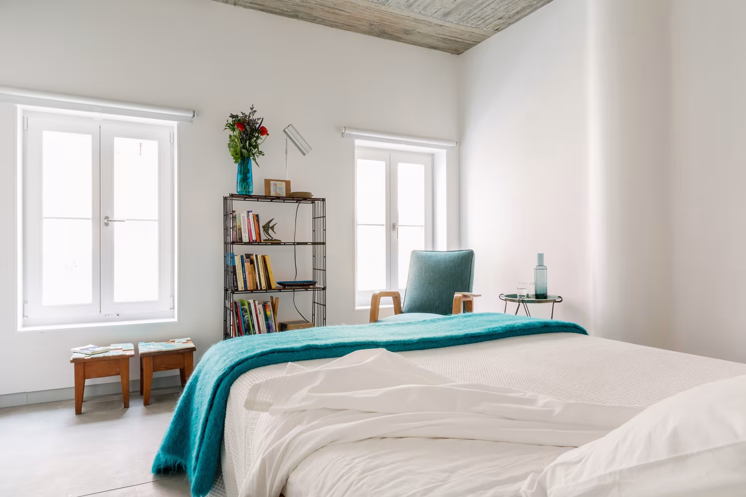 Habitación con cama de manta turquesa, estantería metálica con libros y ventanas blancas en el alojamiento de Lisboa
