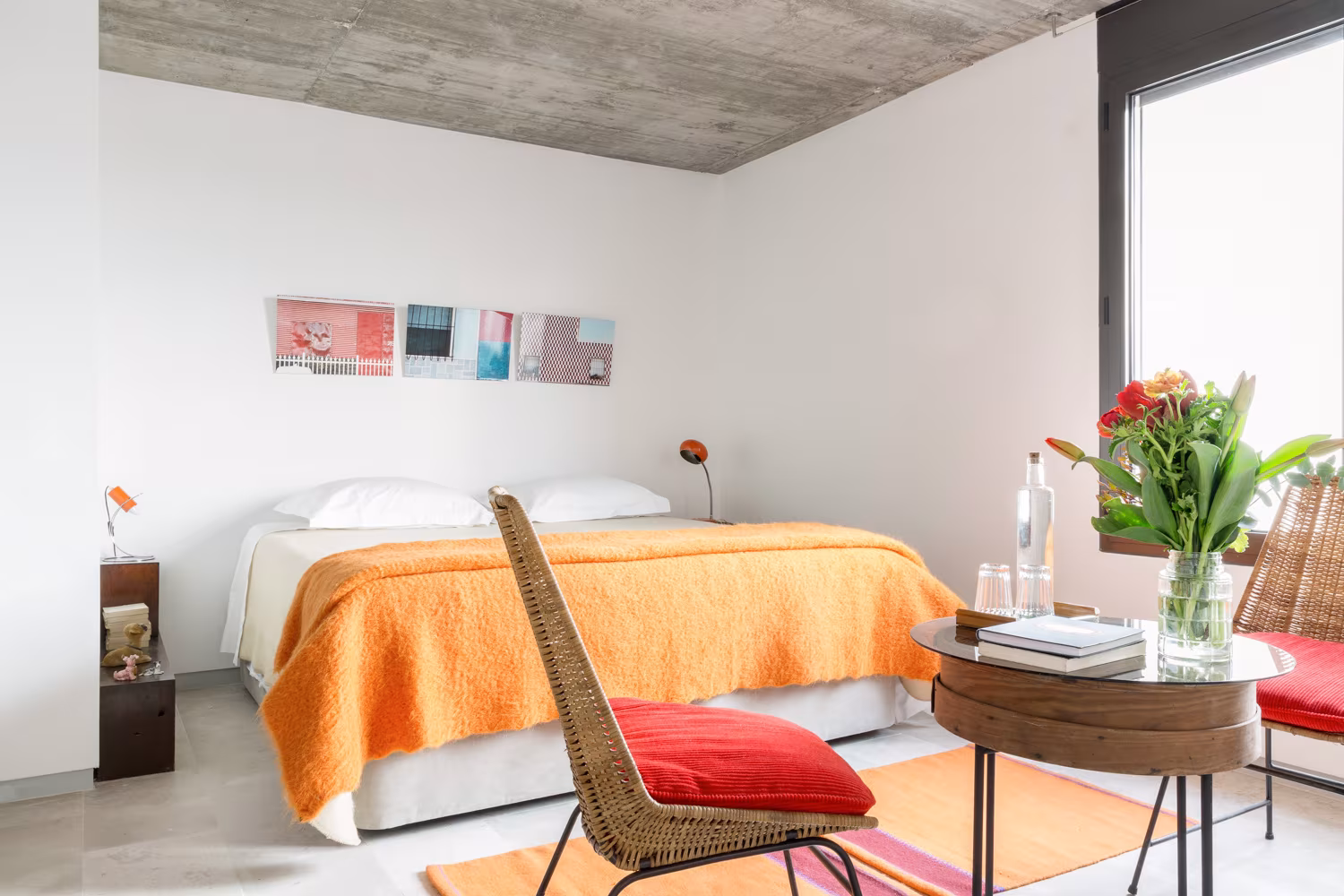Habitación con colcha naranja, butaca de rejilla y mesa auxiliar bajo techo de hormigón en el alojamiento de Lisboa