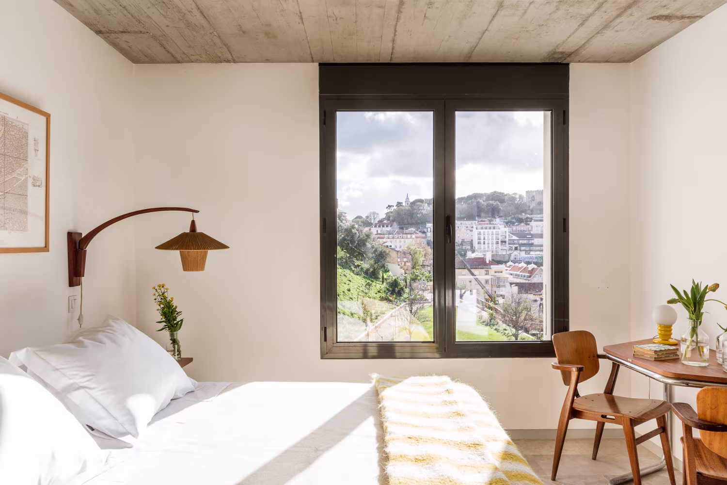 Dormitorio con ventana a los tejados de Lisboa, aplique de mimbre y silla de madera en el alojamiento singular