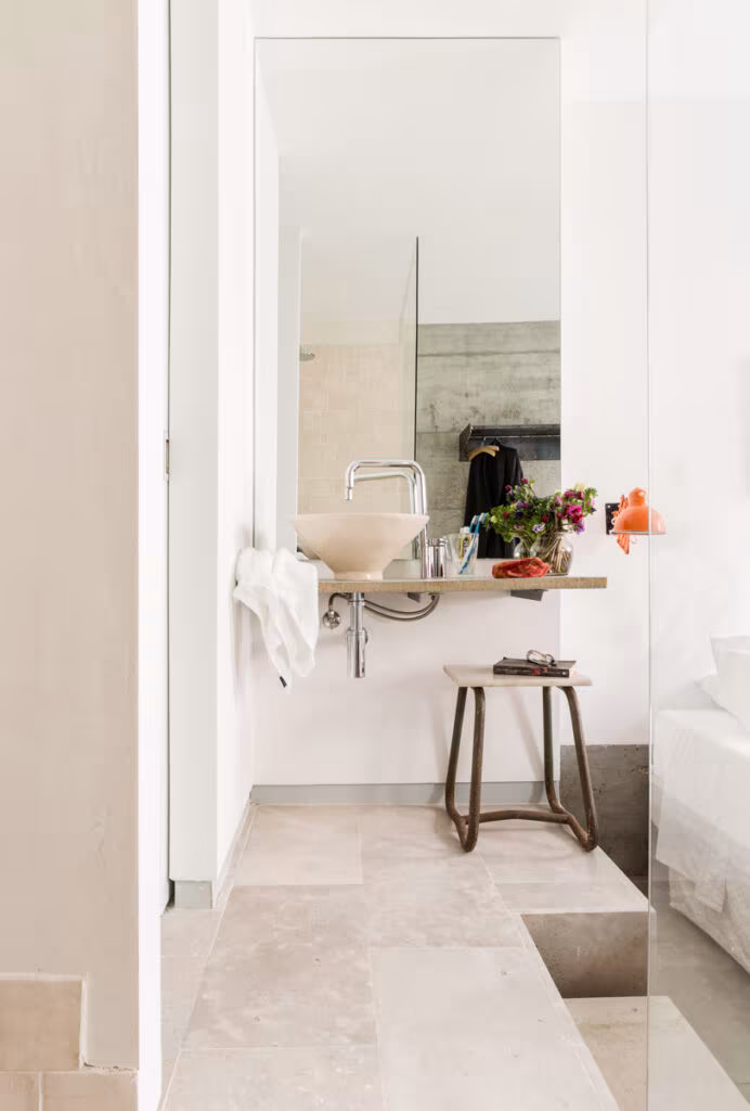 Baño abierto al dormitorio con lavabo sobre repisa, taburete y suelo de piedra clara en el alojamiento de Lisboa