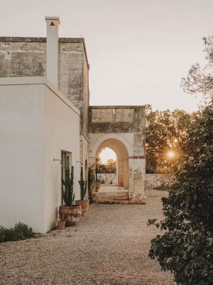 Camino de grava entre muros de estuco blanco y piedra hacia un arco que enmarca el sol en la Casa Casolare Scarani