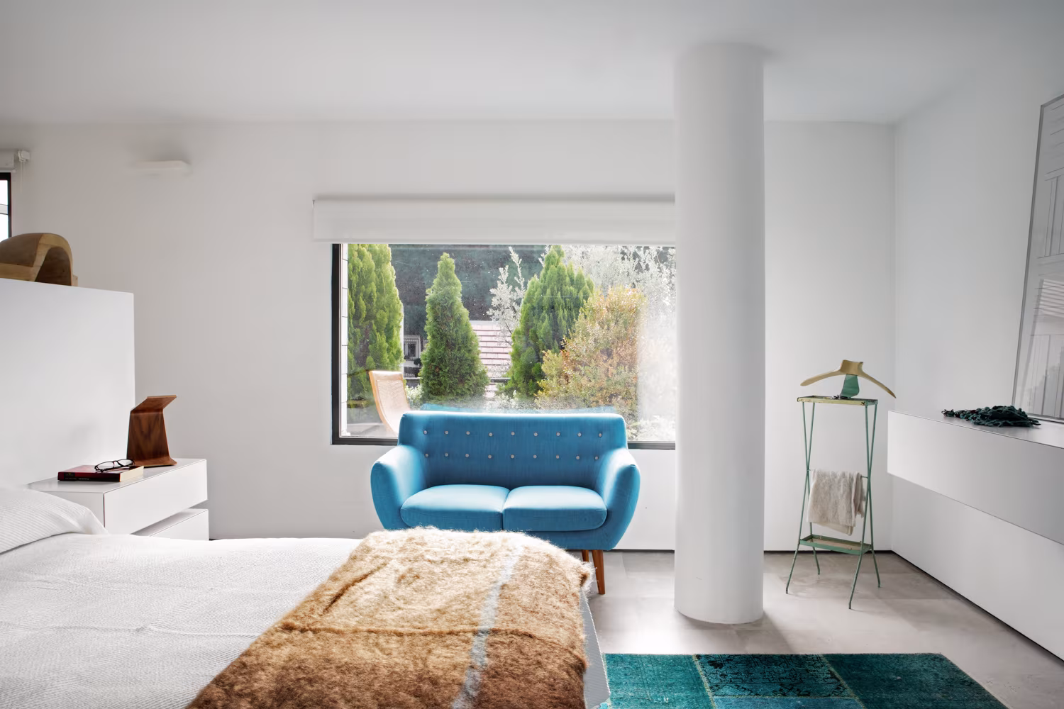 Dormitorio de Casa Oviedo con butaca azul, pilar blanco y ventana al jardín de cipreses