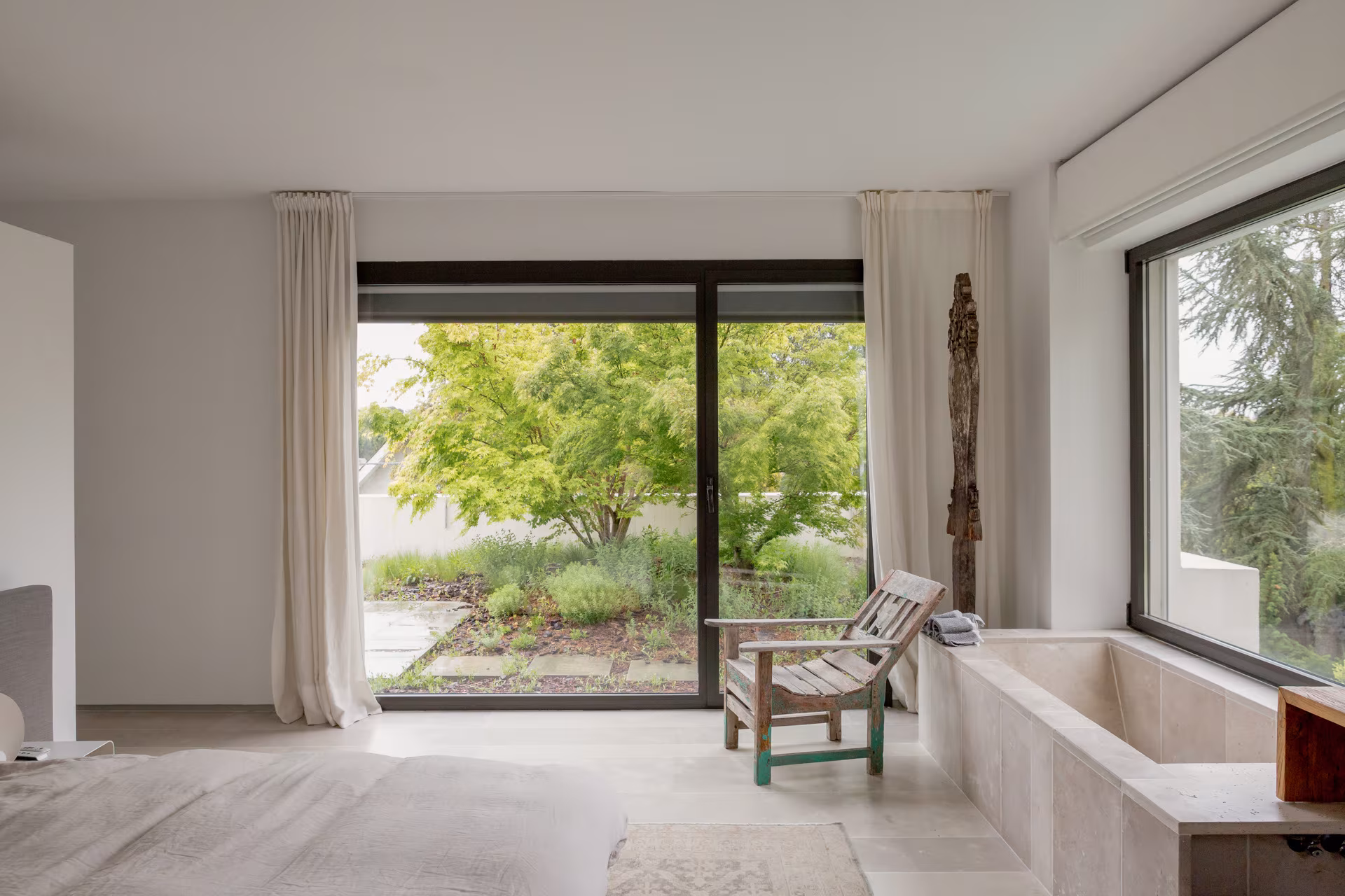 Room with stone bath, timber armchair and large sliding window to the garden with linen curtains, Ábaton studio
