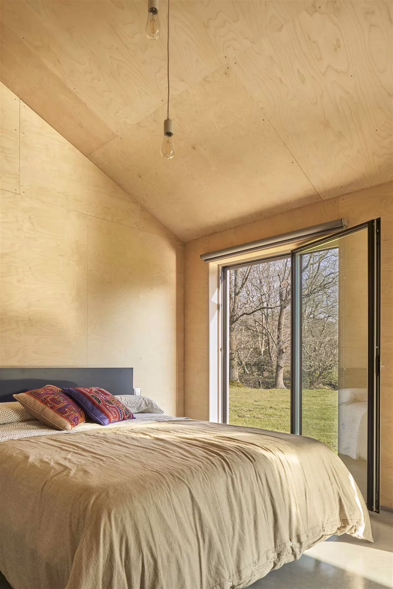 Dormitorio de la casa de Navajeda revestido de madera contrachapada, con ventanal corredero al prado
