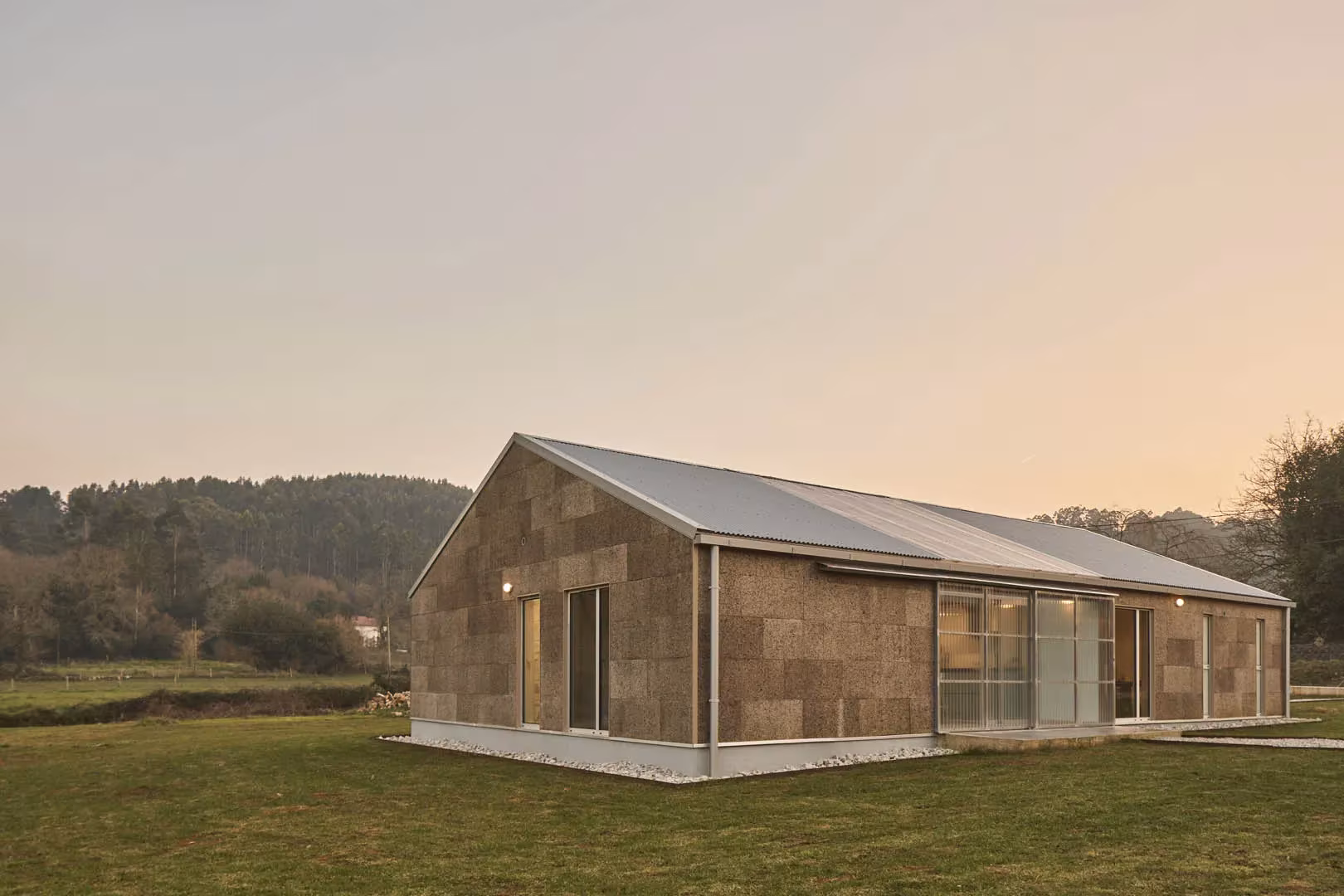 Casa invernadero de Navajeda al atardecer, con volumen alargado de corcho y cubierta a dos aguas en el prado