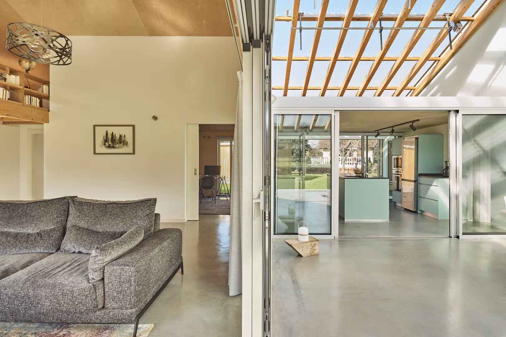 Salón de la casa de Navajeda con sofá gris y cocina al fondo, bajo la cubierta traslúcida de vigas de madera