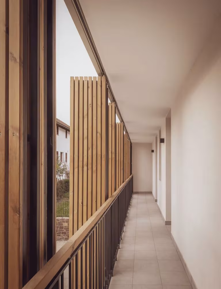 Pasillo de acceso con barandilla y celosía de listones de madera abierto al exterior en las viviendas de CLT