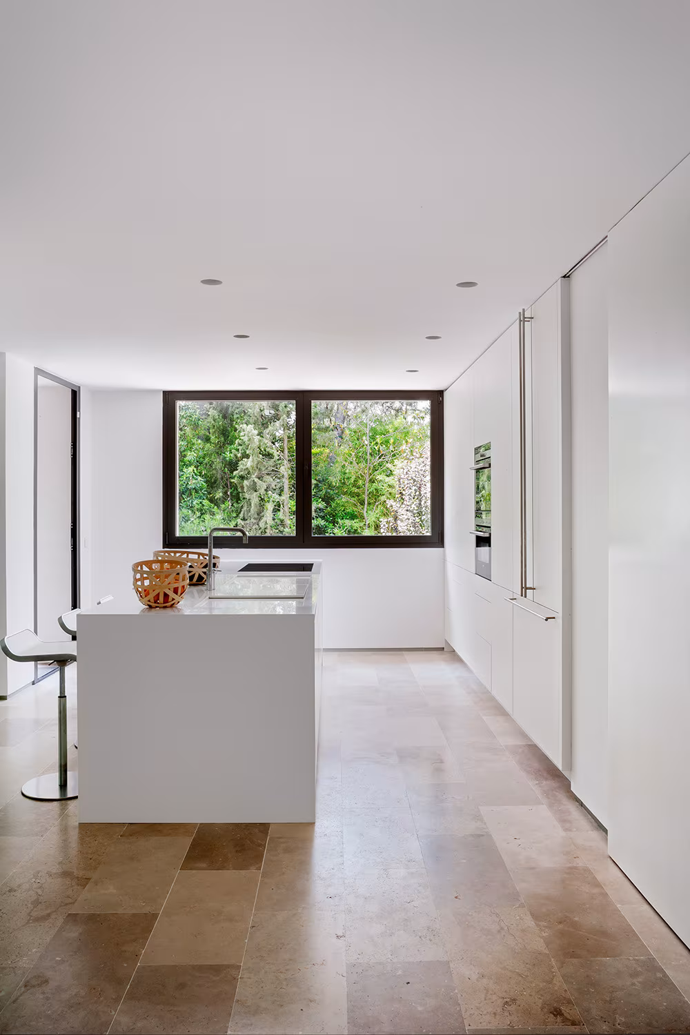 La Moraleja-ABATON-9 White kitchen with island and stone floor, a window to the trees at the end, La Moraleja refurbishment