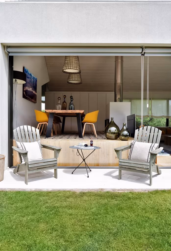 Porch with timber armchairs and a yellow-chaired dining area behind the open glazing, Proyecto XY