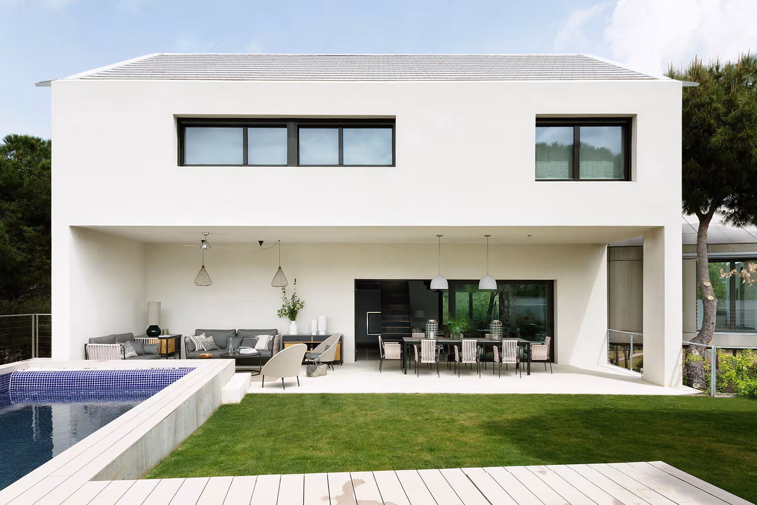 White rear façade of Proyecto XY with porch, outdoor dining, lawn and blue-edged pool