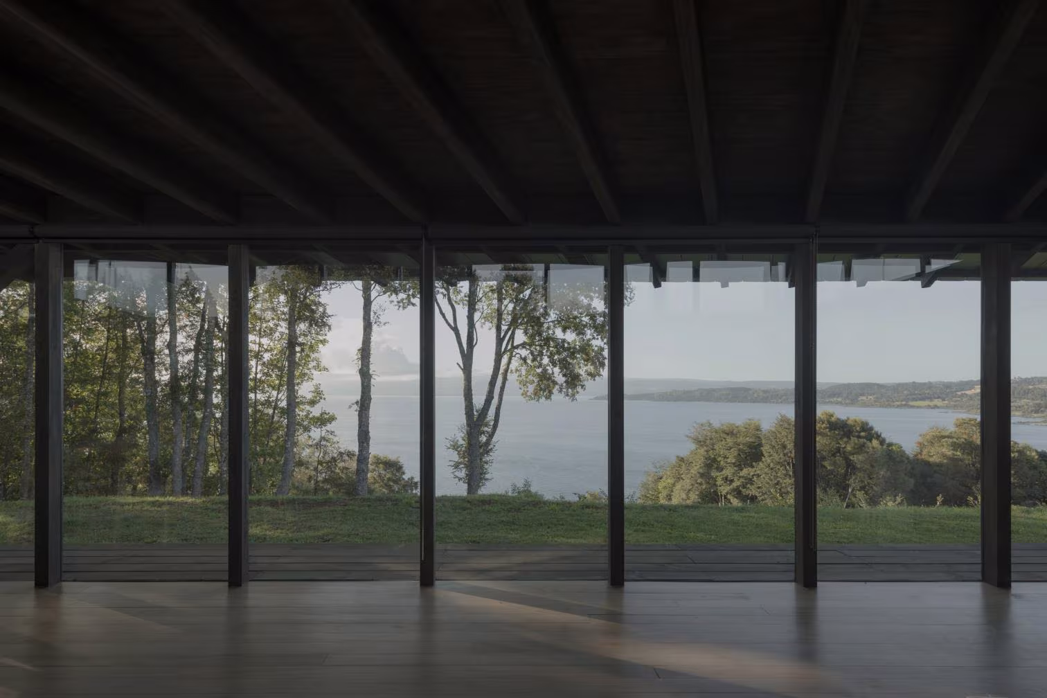 Salón acristalado de Casa Puerto Nuevo con vistas al lago entre los árboles y estructura de madera oscura