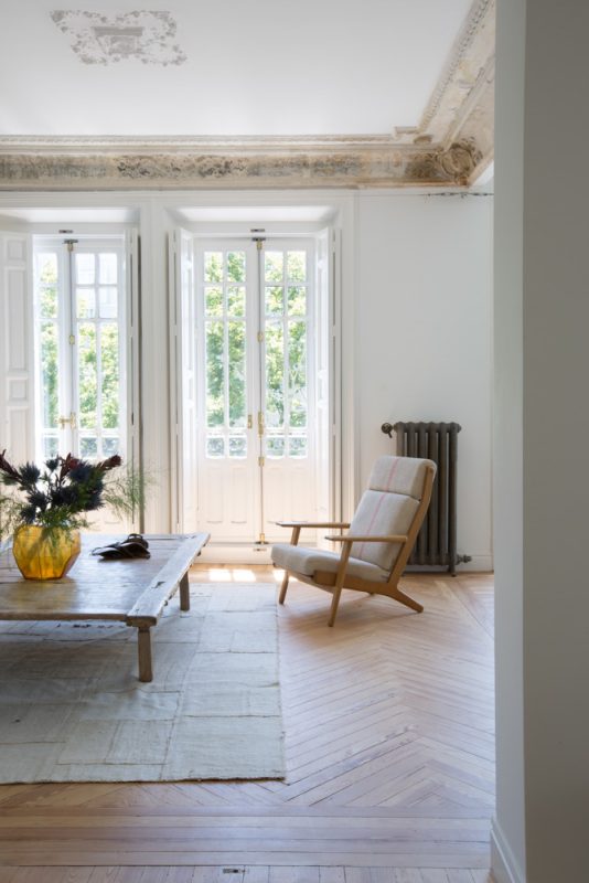 ABATON-reforma-integral-en-sagasta-12 Salón luminoso de estilo clásico renovado con molduras ornamentales en el techo, ventanales franceses con contraventanas blancas, suelo de madera en espiga y mobiliario minimalista con mesa rústica de madera y sillón escandinavo tapizado