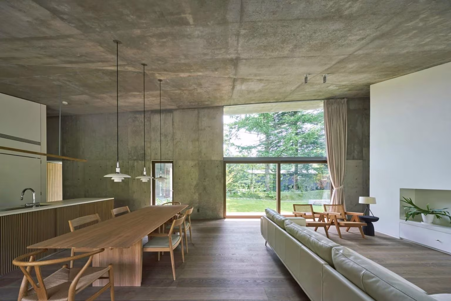 Timber kitchen with a long dining table and pendant lamps facing a large window in the Symbiotic House