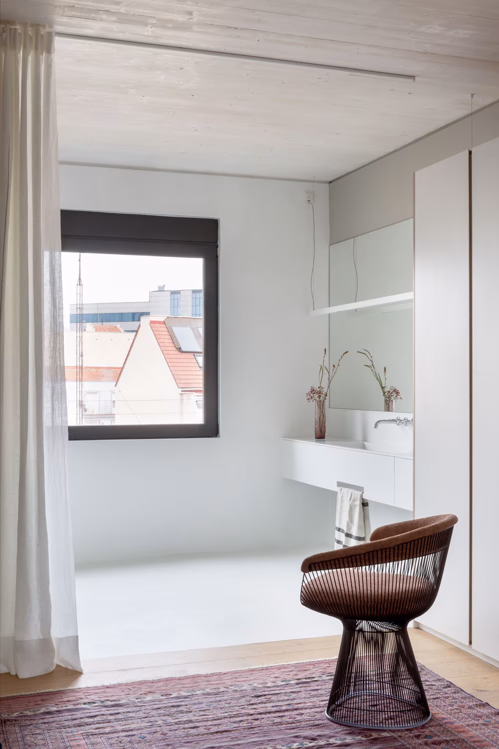 Dormitorio de ARV8 con cortina blanca, ventana de marco negro y butaca de rejilla metálica sobre alfombra