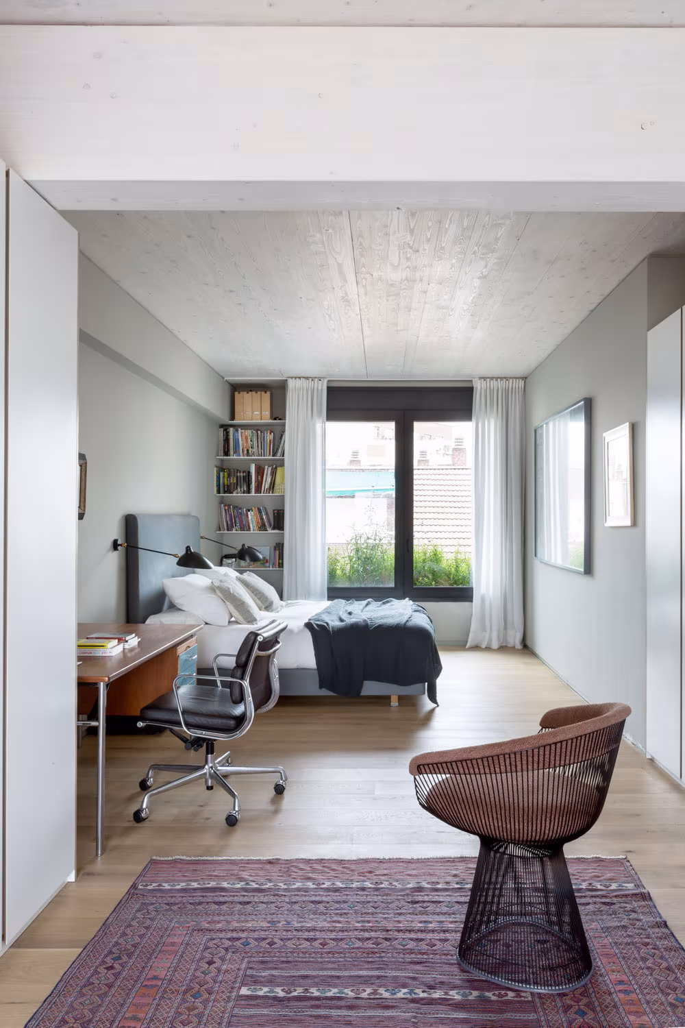 Dormitorio de ARV8 con escritorio, estantería y butaca junto a la ventana, bajo un techo de madera contralaminada