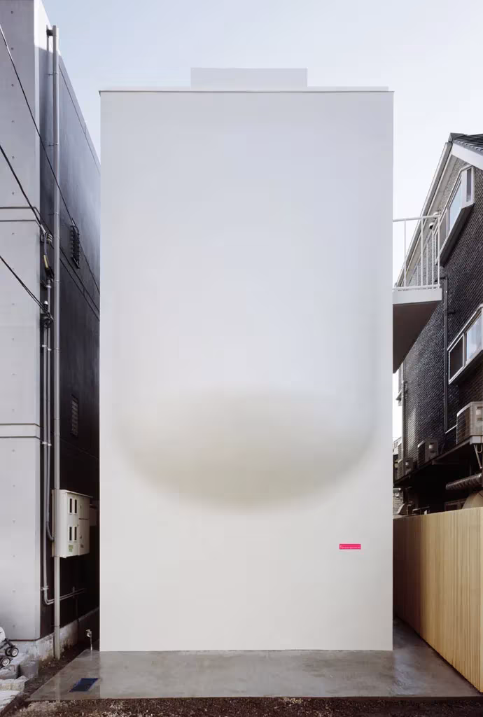 Fachada ciega y blanca de la House SH de Hiroshi Nakamura, con su base curva y un rótulo rojo en el lateral
