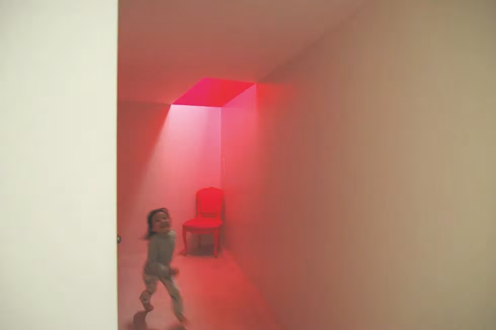 Pequeño espacio de la House SH bañado en luz roja por un lucernario, con una niña y una silla roja