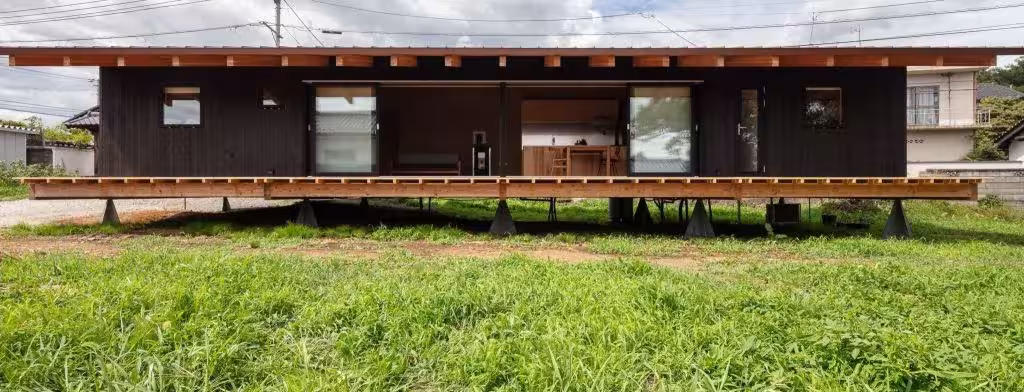 Casa levitante de fachada de madera oscura elevada sobre apoyos en un prado, con una larga terraza corrida