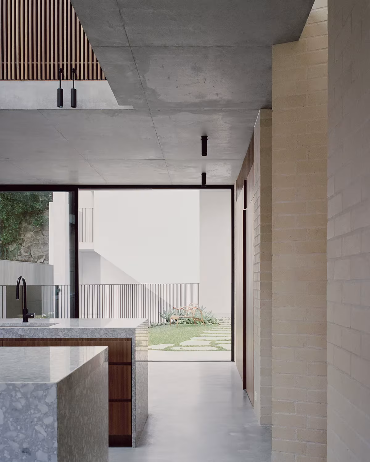 Cocina de la Bronte House con isla de terrazo, ladrillo visto y ventana al jardín bajo un techo de hormigón