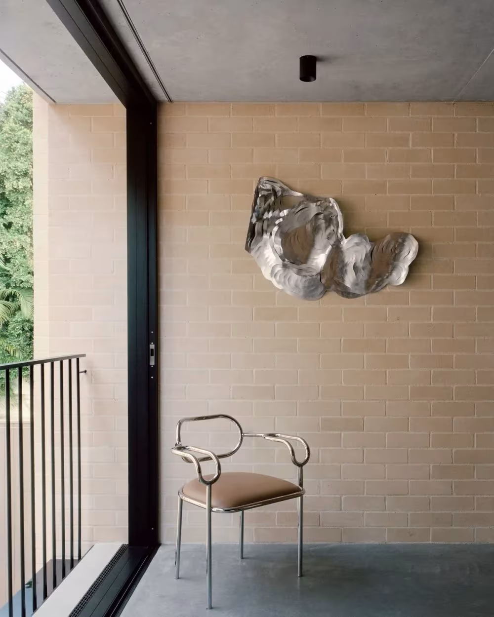 Ventana corredera y silla de metal ante un muro de ladrillo con una escultura plateada, en la Bronte House
