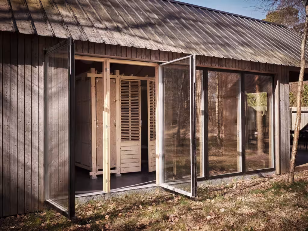 Cabaña de madera oscura con puertas de vidrio abiertas y celosías interiores de tablillas en un entorno boscoso