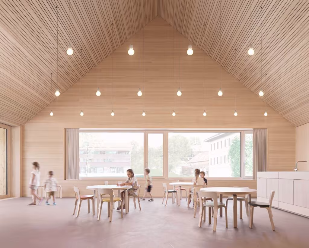 Aula con cubierta a dos aguas de listones de madera y niños sentados a mesas redondas en la Casa de los niños