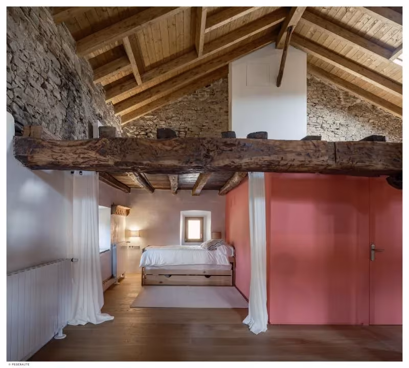 Dormitorio bajo la cubierta de madera de Garaitia Etxea, con panel rojo, cortinas y vigas antiguas a la vista
