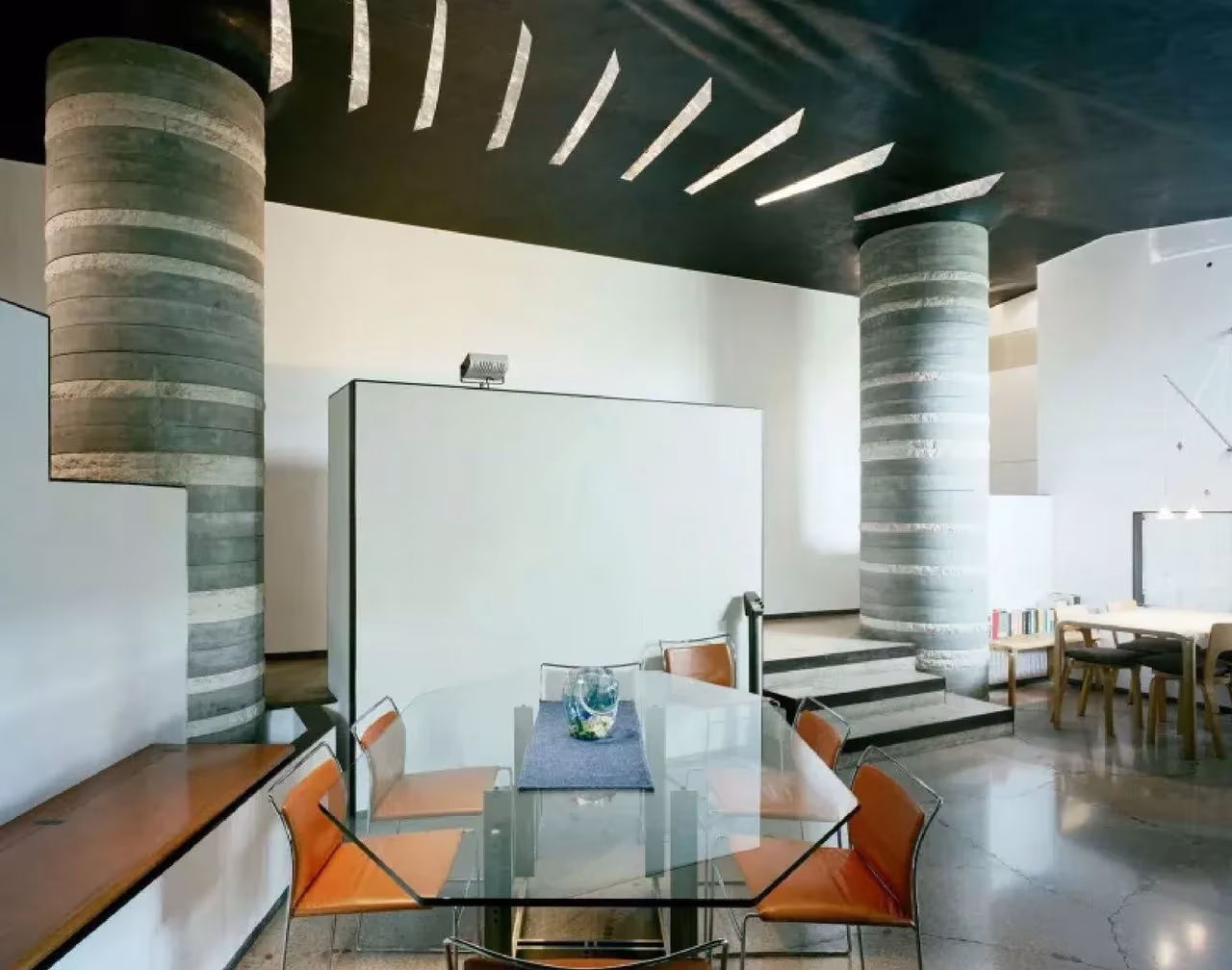 Salón-comedor de la Villa Ottolenghi con mesa de cristal, columnas de hormigón anilladas y techo oscuro con lamas de luz