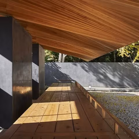 Banco corrido de madera bajo la cubierta de lamas del pabellón, con luz y sombra sobre la grava