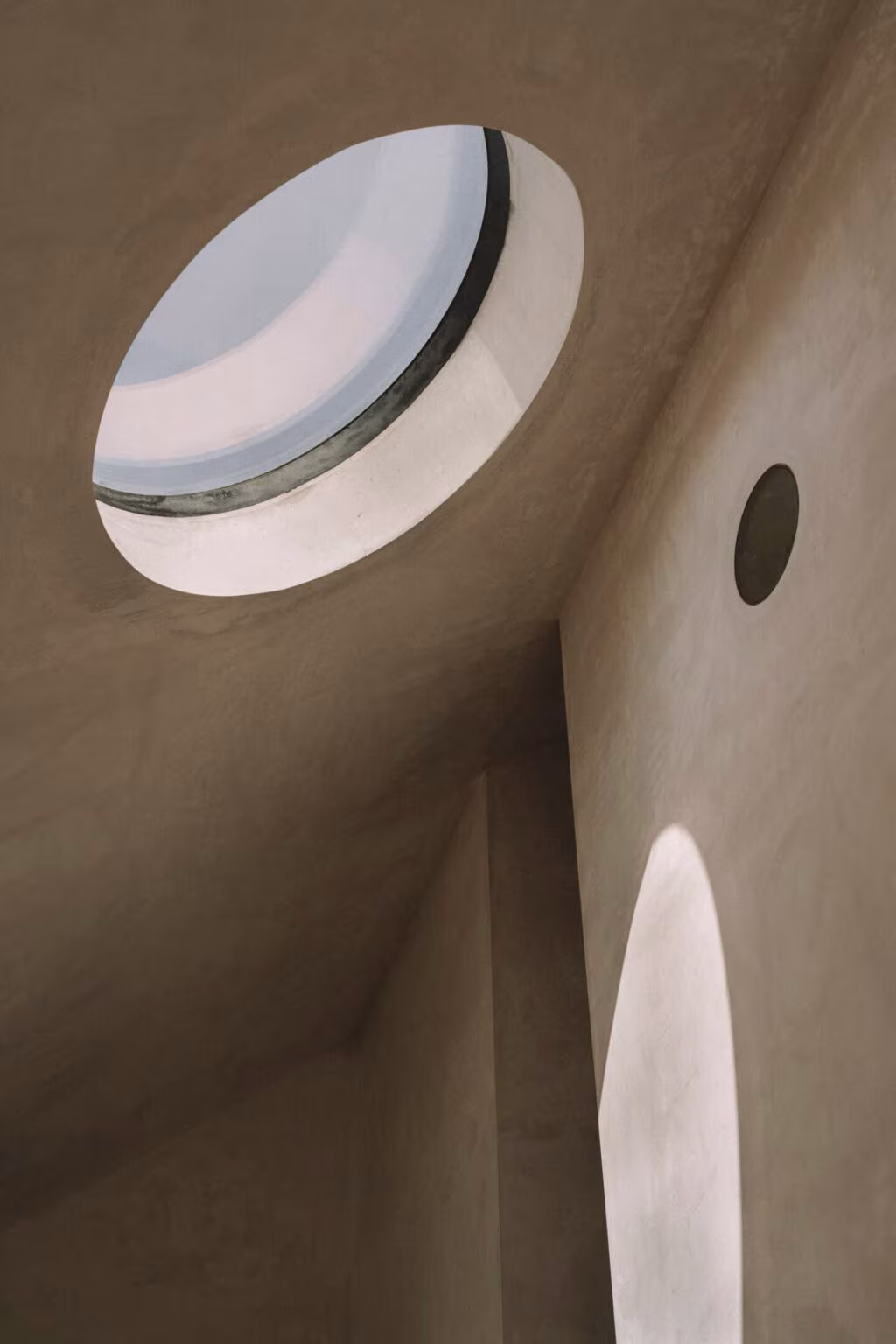 Circular rooflight in the sloping plaster ceiling of Casa Plaj, with a patch of sun on the wall
